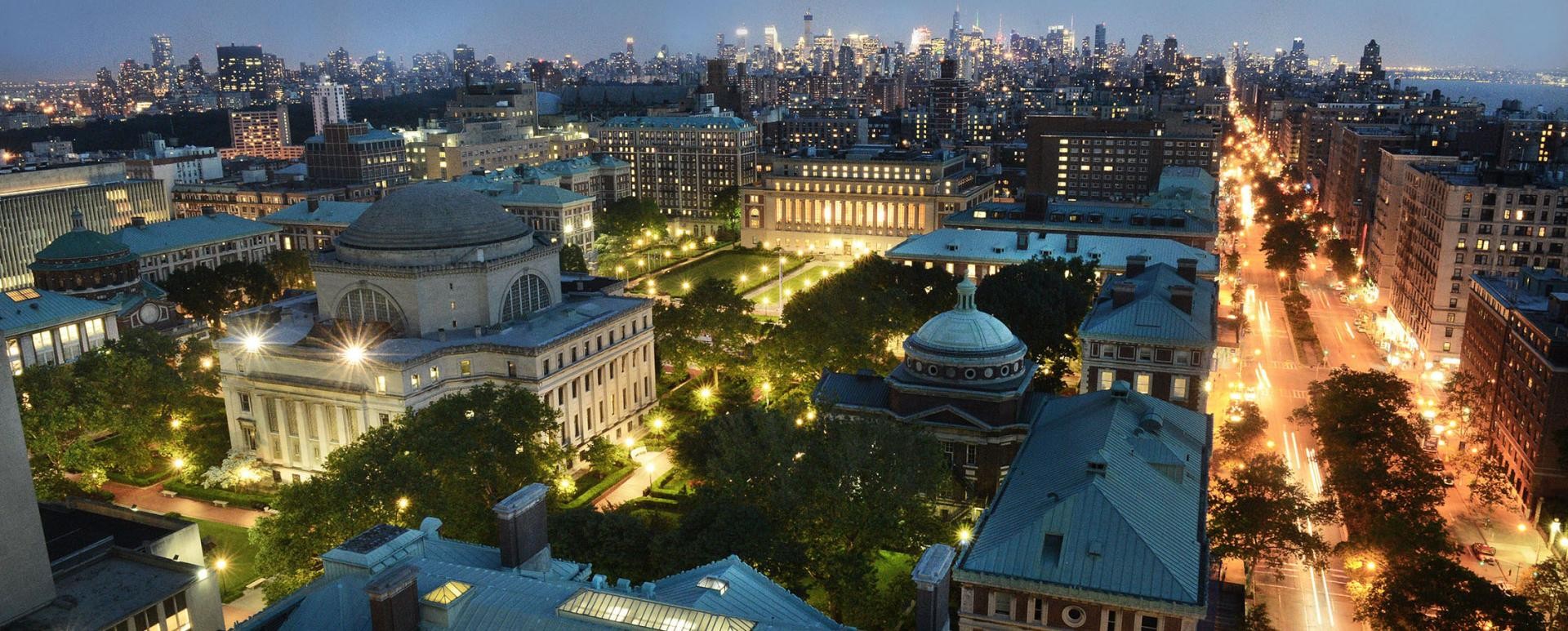 Angled view of Columbia's Morningside Heights campus at night with the Manhattan skyline in the background